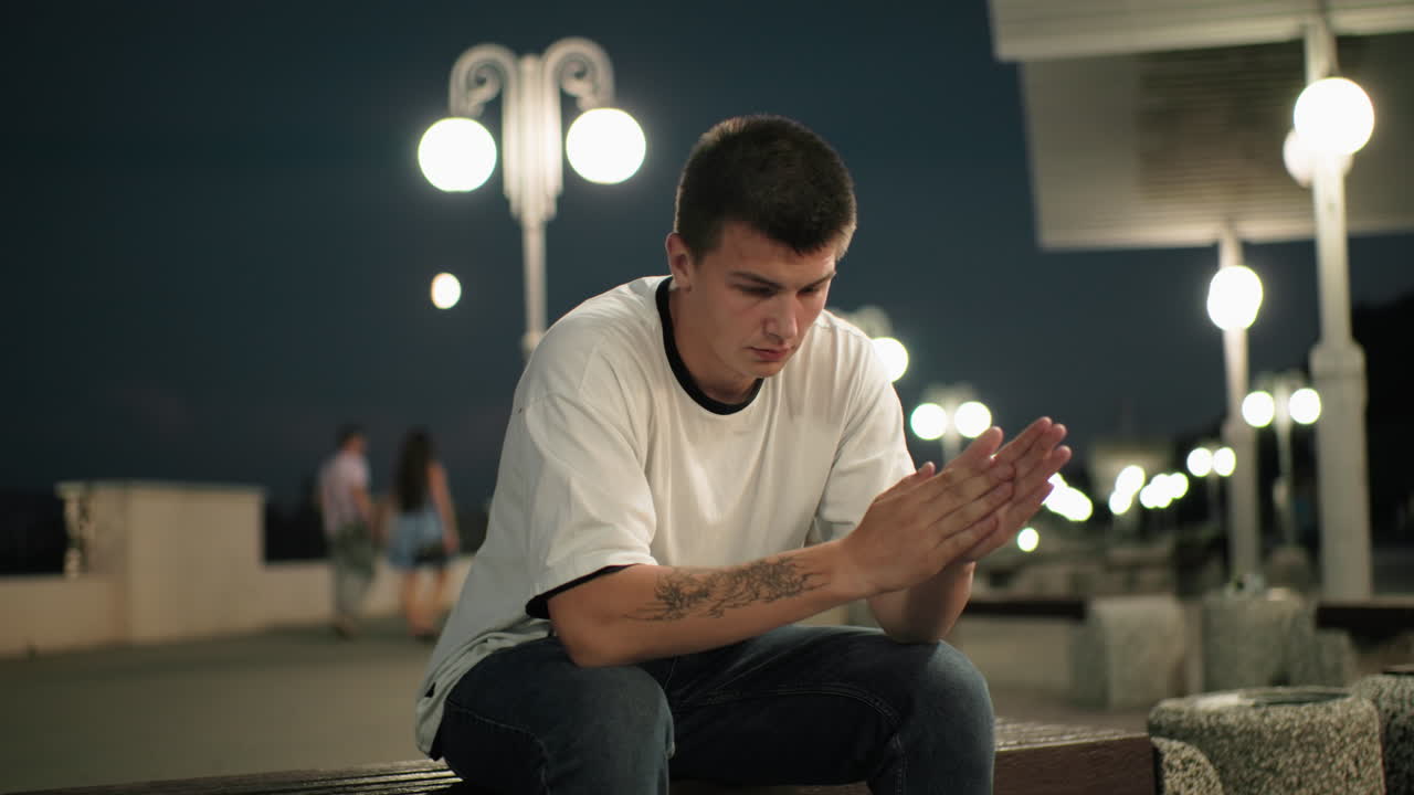 문신한 손을 가진 사려 깊은 남자가 저녁에 벤치에 앉아, 깊은 생각에 빠져, 거리 조명과 흐릿한 커플이 지나가는 도시 배경