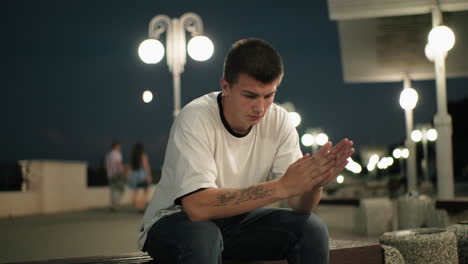 hombre contemplativo con mano tatuada sentado en un banco al anochecer, profundamente en pensamiento, fondo urbano con farolas y parejas borrosas que pasan