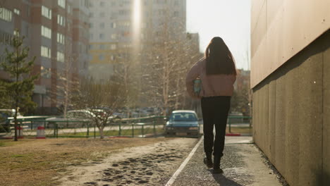 back shot of a girl in a peach jacket and black pants, holding a turquoise skateboard, walking along a sunlit pathway beside a building , with trees, cars, and apartment in the background