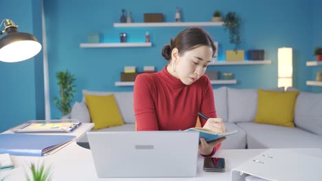 asian young woman working from home thoughtfully taking notes and working from laptop.