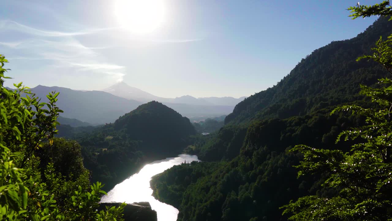 Handheld of river surrounded by mountains covered in dense woods, Villarrica volcano in background, El Leon viewpoint, Chile