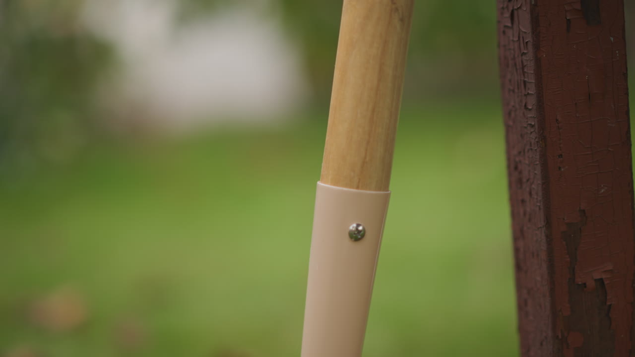 Primer plano del mango de una pala apoyado en un poste de cerca, bokeh de césped verde suave, virola de metal, textura de madera con sutiles gotas de lluvia, hojas de otoño desenfocadas, bodegón de jardín tranquilo, poca profundidad de campo cinematográfica