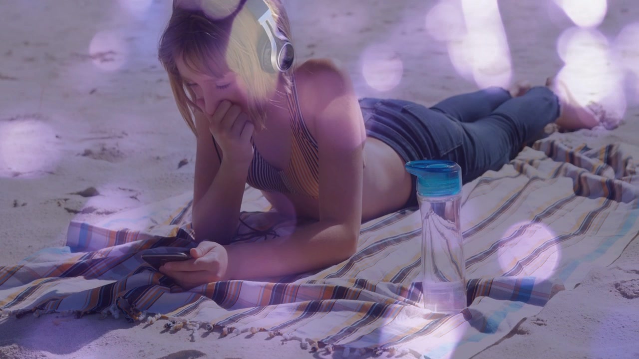 Ragazza sdraiata su un tappetino a strisce in spiaggia con le cuffie, che tocca un tablet attivando un bokeh viola per la tecnologia.