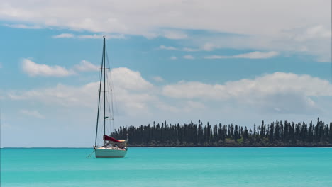 un velero anclado se desplaza en el océano azul celeste con una sobrecarga de nubes y pinos caledonianos en el fondo - lapso de tiempo