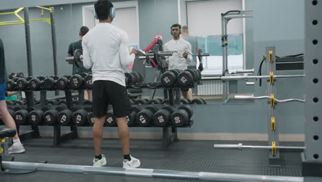 mirror reflecting man lifting chrome barbell while others pass by and work out in gym, showing dumbbell racks, rubber flooring, metal bars and active workout session in modern fitness facility