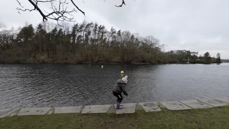 Ein-Kleiner-Junge-Hat-Spaß-Daran,-An-Einem-Kalten-Wintertag-Mit-Hut-Und-Mantel-Am-Ufer-Eines-Sees-Entlangzulaufen