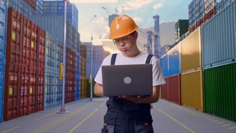 trabajador asiático con gafas de protección y casco de seguridad usando una computadora portátil y con dolor de cabeza mientras está de pie en el almacén del patio de contenedores