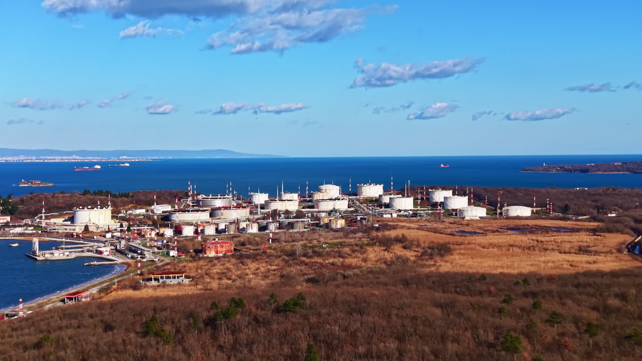 Complexo industrial perto da costa com tanques de armazenamento e uma grande massa de água ao fundo durante o dia.