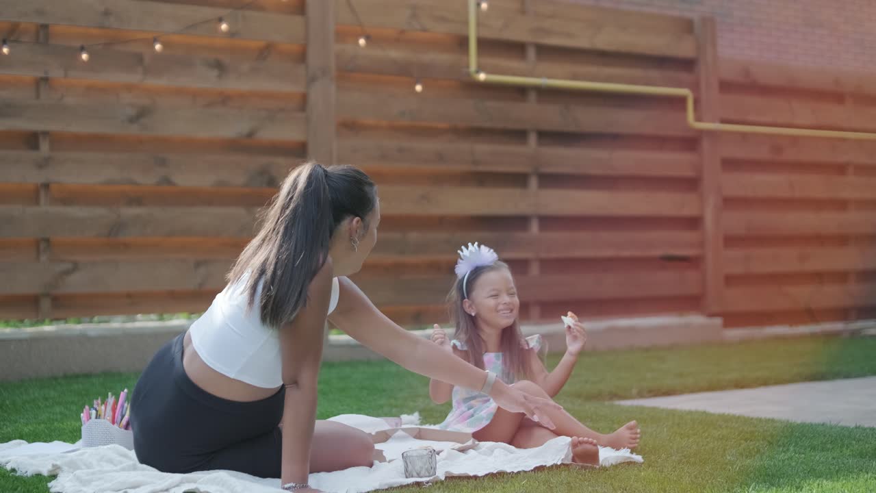 엄마와 어린 딸이 뒷마당에서 담요 위에 앉아 함께 그림을 그리고 있습니다. 야외 장면은 평화로운 가족 순간을 포착하여 창의성과 유대를 촉진합니다.