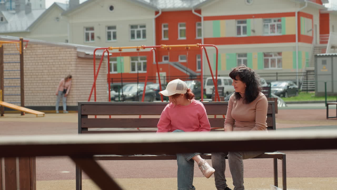 Caucasian Woman And Teen On Playground Bench During Outreach Chat, Colorful Apartment Complex Behind, Rabbit Resting Nearby, Empathetic Gestures And Quiet Support, Volunteer Style Interaction