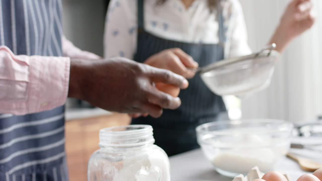 Biracial couple wearing aprons and baking in kitchen, slow motion