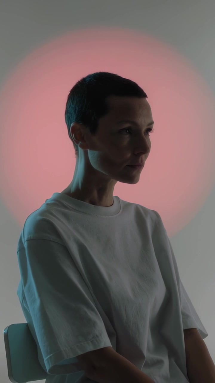 Video verticale: Donna che muove la testa e gli occhi in studio, con una maglietta bianca con alone rosa e bordo verde acqua.