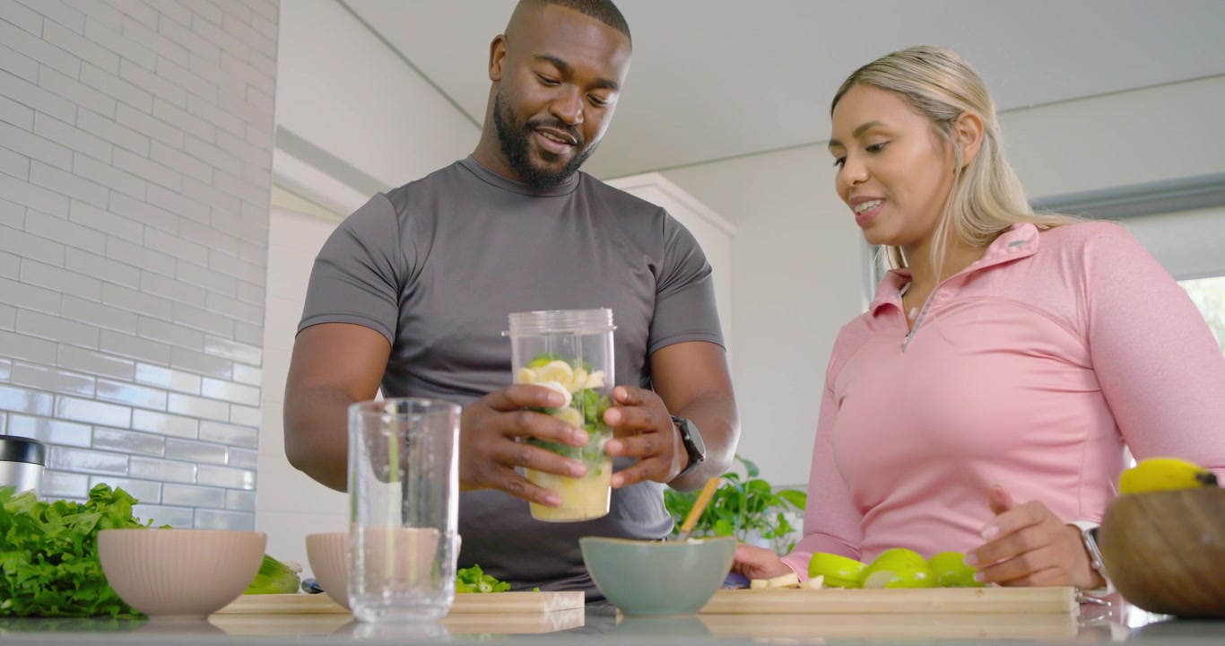 Casal afro-americano em forma, mulher adicionando vegetais no liquidificador no balcão para uma bebida, homem segurando uma xícara.