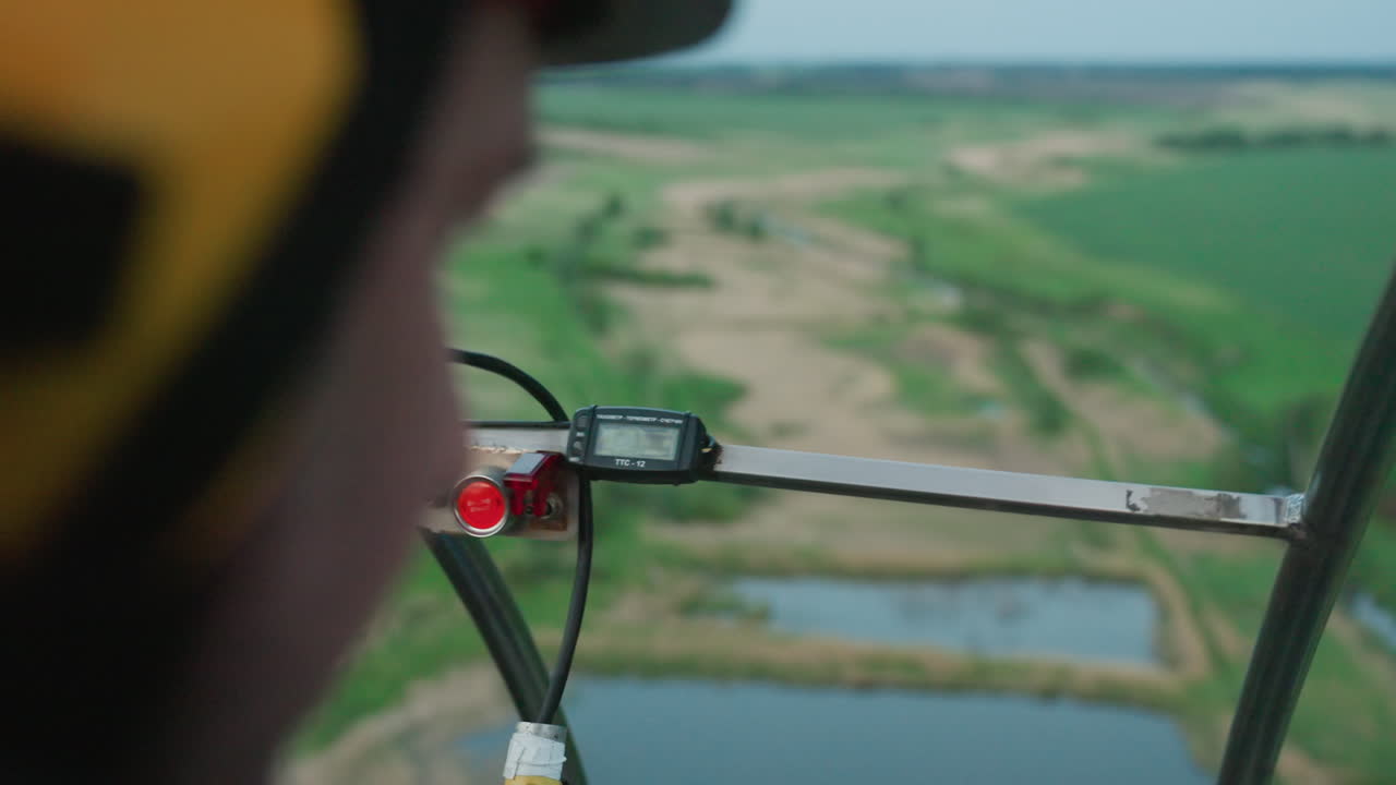 overhead cockpit view showing pilot operating camera rig filming expansive green fields with ponds and winding dirt paths from aircraft under soft daylight capturing aerial exploration