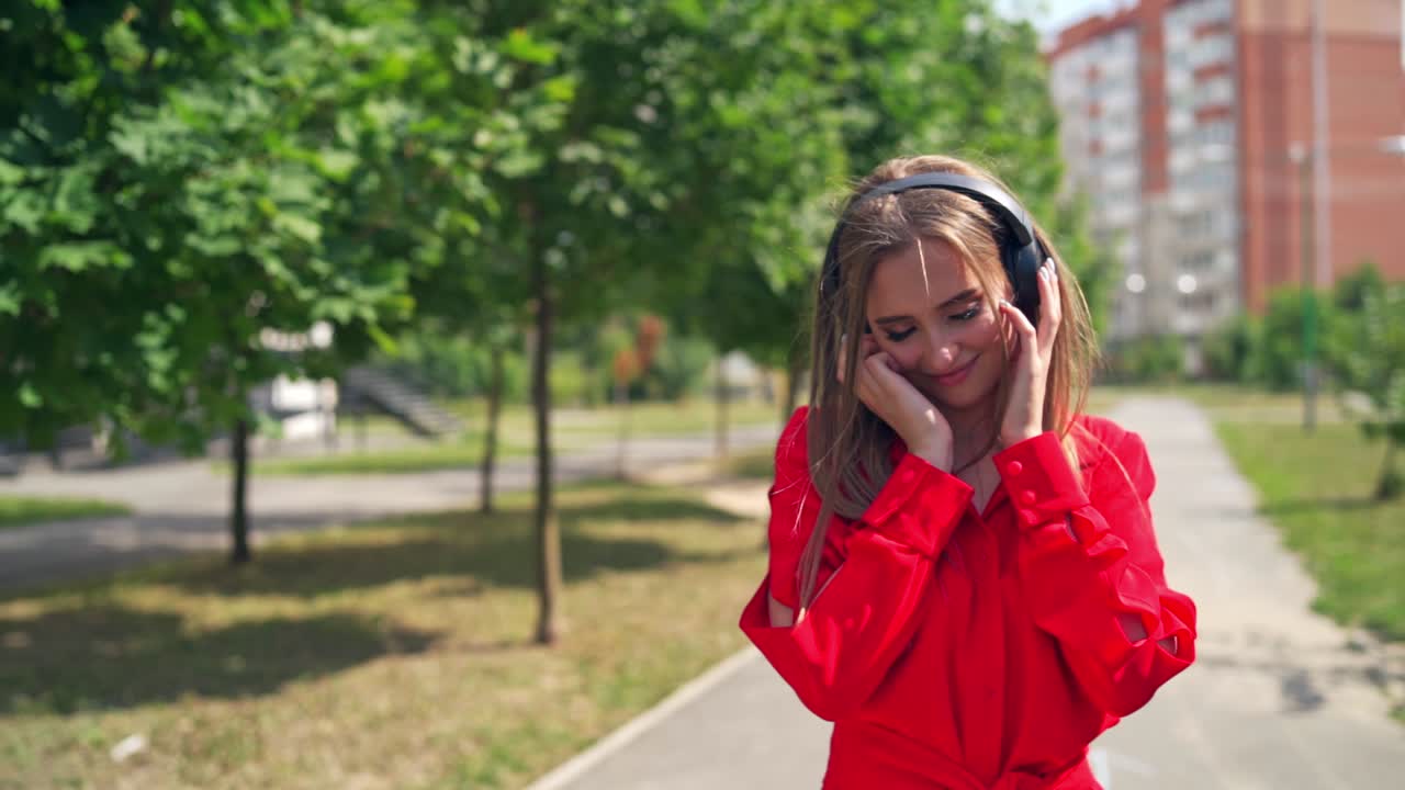 Lovely girl listening to music outdoors. Beautiful young woman with black headphones standing in the street and enjoys music on the city background in summer.