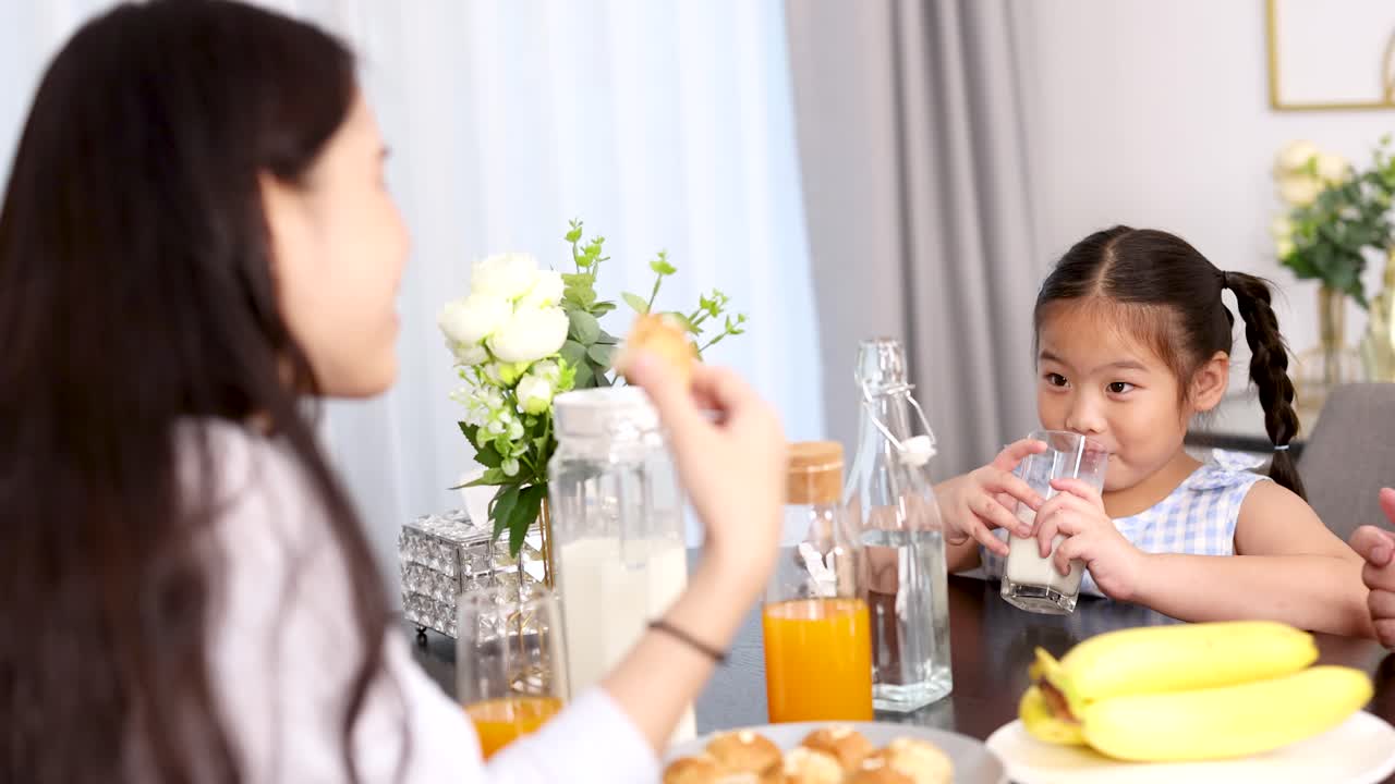 Young Asian girl drinks milk at breakfast table with family, surrounded by fruit and sunlight