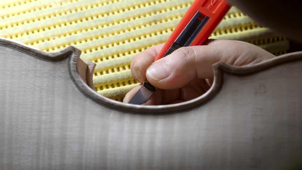 Close-up shows a luthier carving the purfling channel on a spruce violin top plate with a sharp utility knife, demonstrating traditional attention to acoustic and aesthetic detail