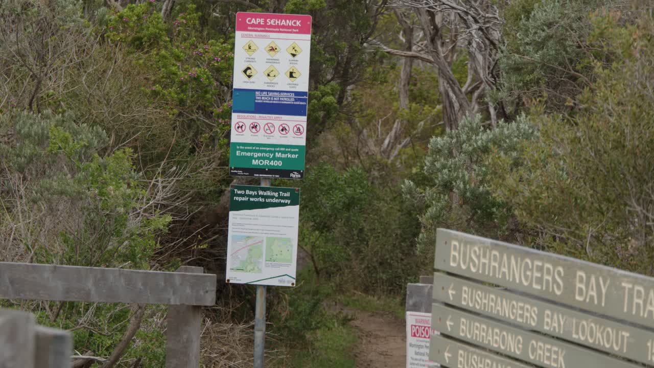 Camera moves toward Bushrangers Bay Track sign at trail entrance, overcast daylight, natural surroundings