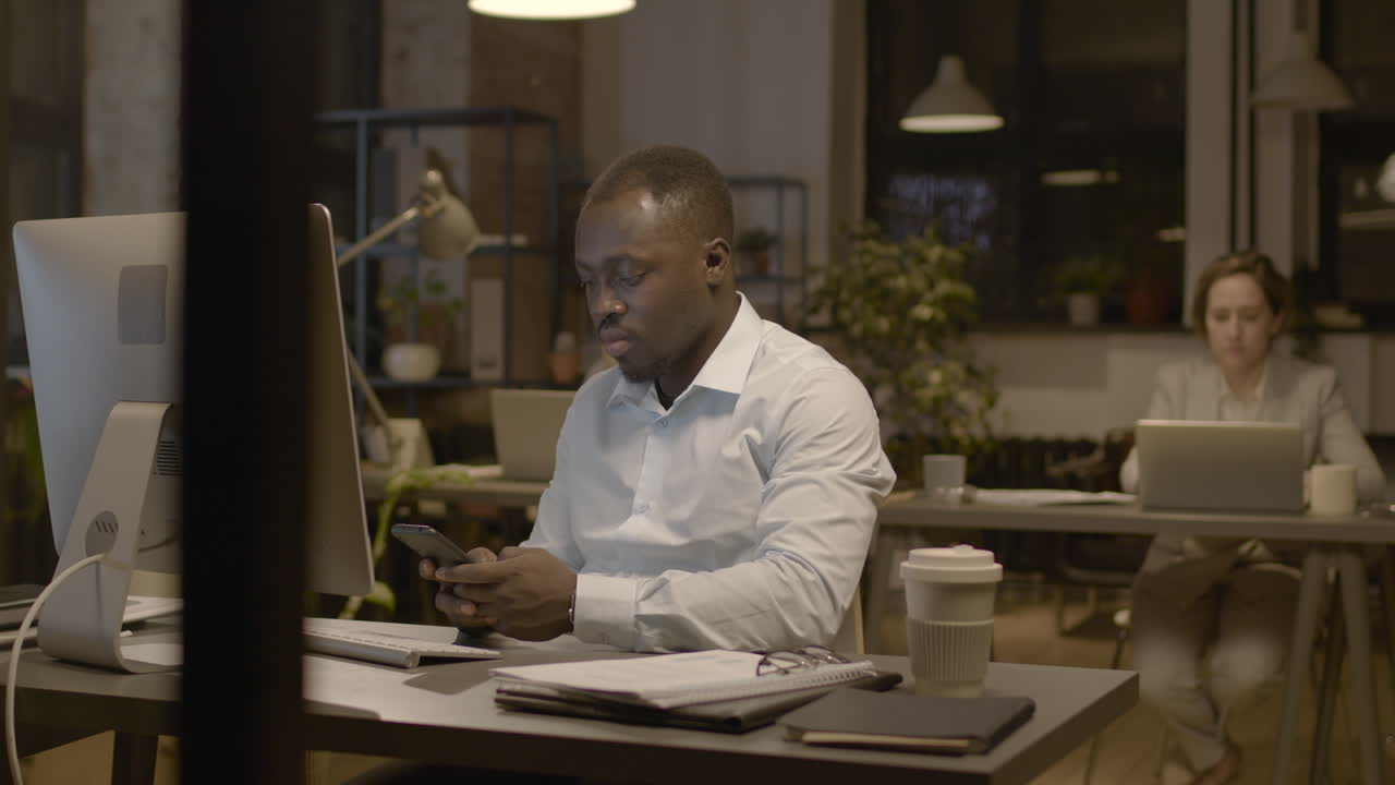 American Man Texting On Smartphone And Woman Employee Working On Computer In The Office