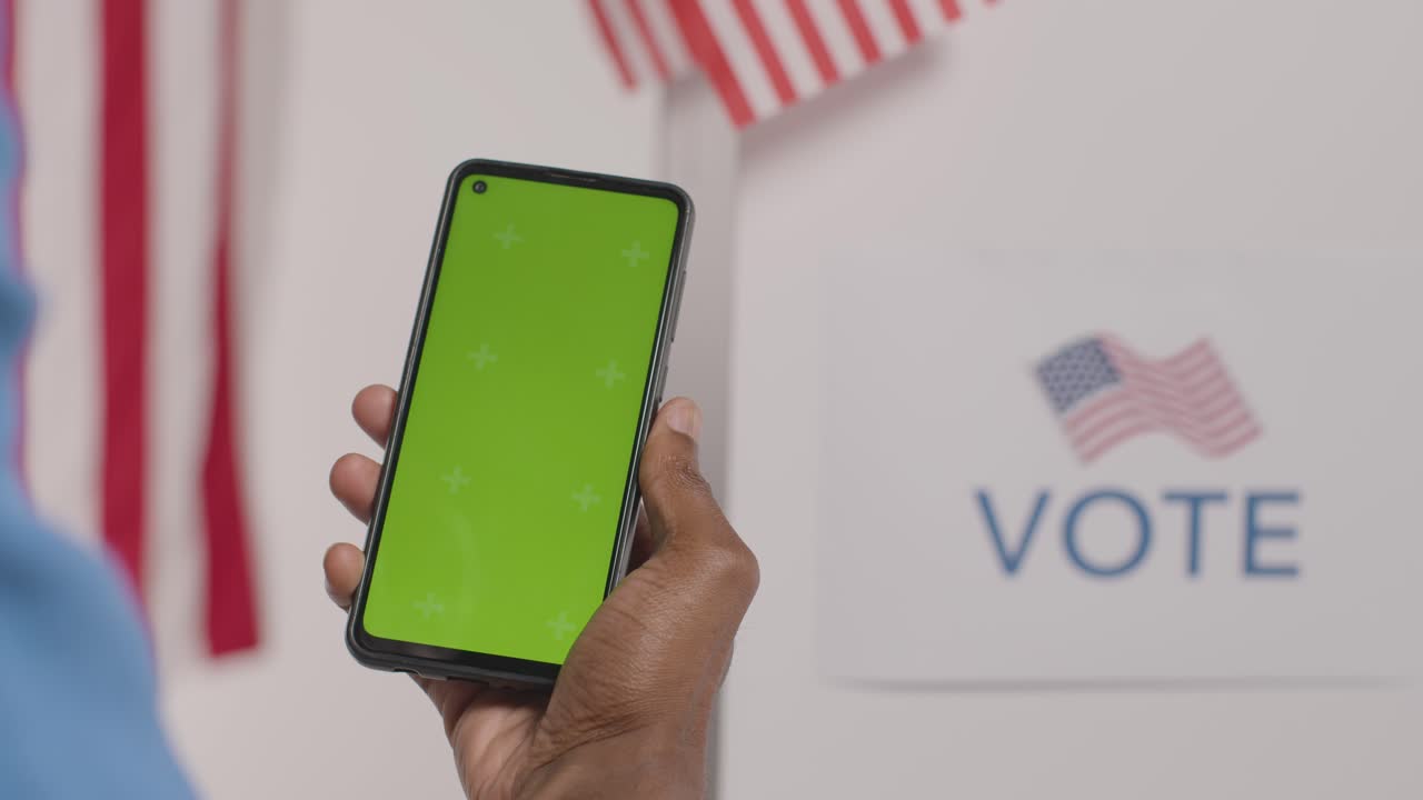primer plano de un hombre con un teléfono móvil de pantalla verde frente a una urna electoral en las elecciones estadounidenses 1