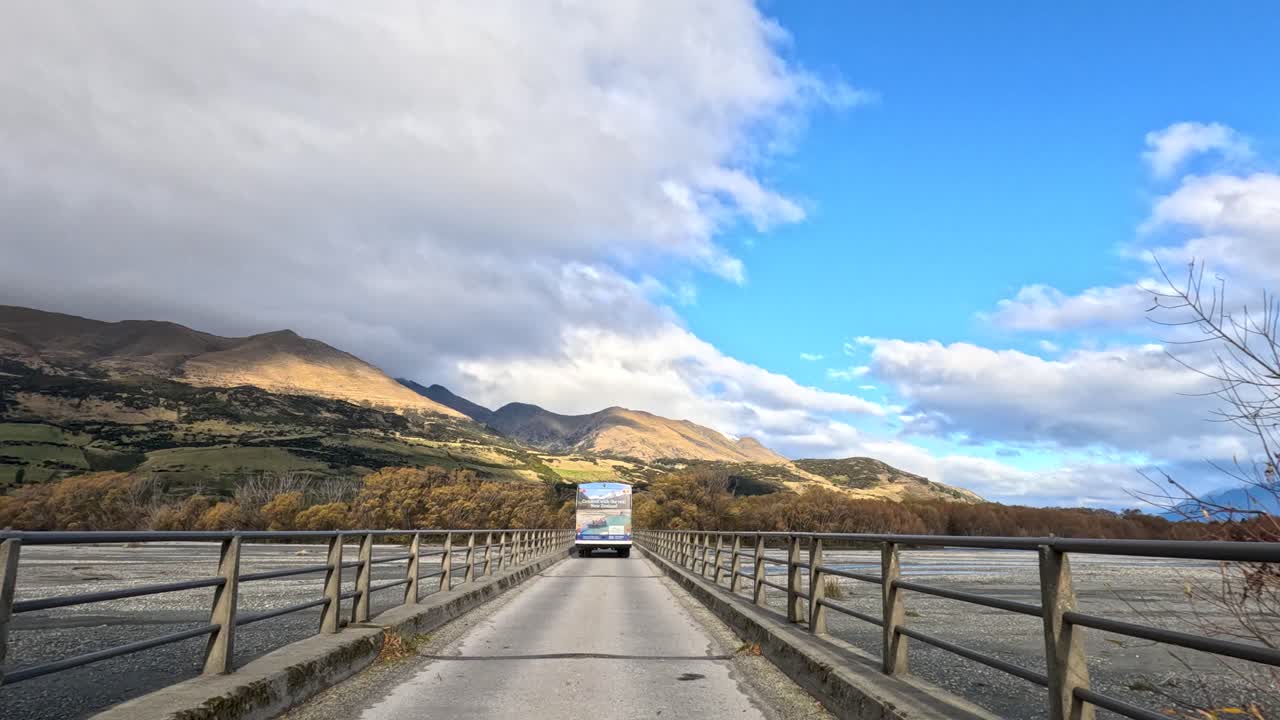 Vehicle drives along tree-lined road and bridge, approaching mountains under bright daylight and blue sky