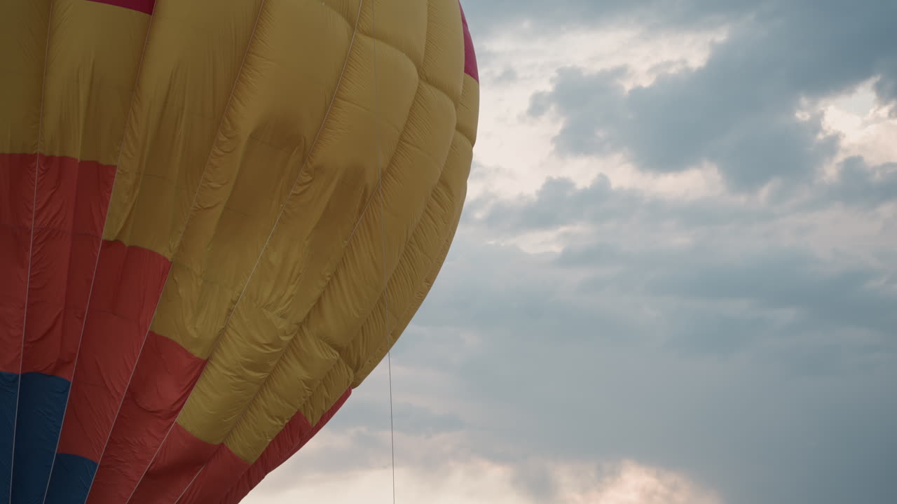 large colorful hot air balloon envelope inflating and rising against cloudy sky at dawn over lush green pasture during adventure flight preparation with vivid fabric panels filling with heated air
