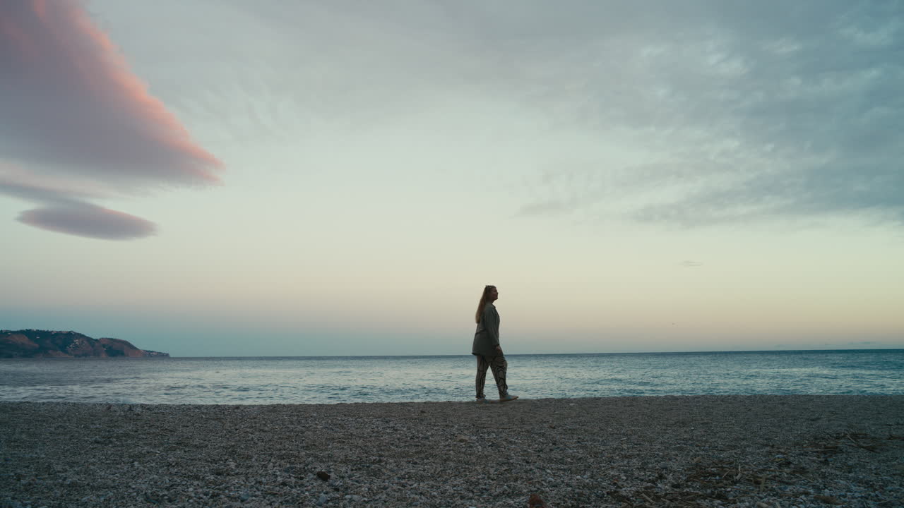 Bredt slow motion-skud af en kvinde, der går alene langs en stenstrand ved solnedgang, hvor det rolige hav og den pastelfarvede himmel skaber en fredfyldt kyststemning.