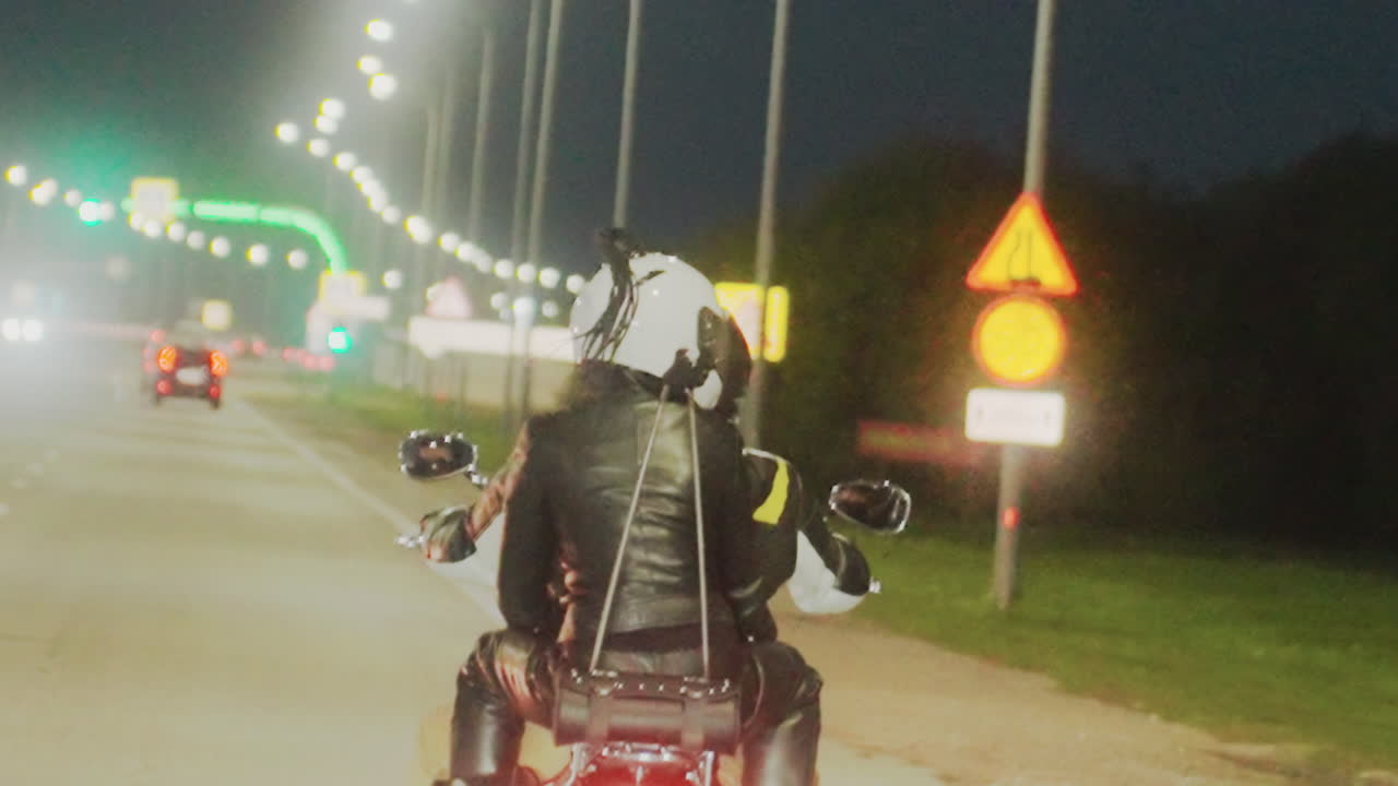 Back view of motorcycle rider in helmet and leather jacket overtaking another biker on illuminated highway, tail light glowing, moving toward city under night sky with visible traffic