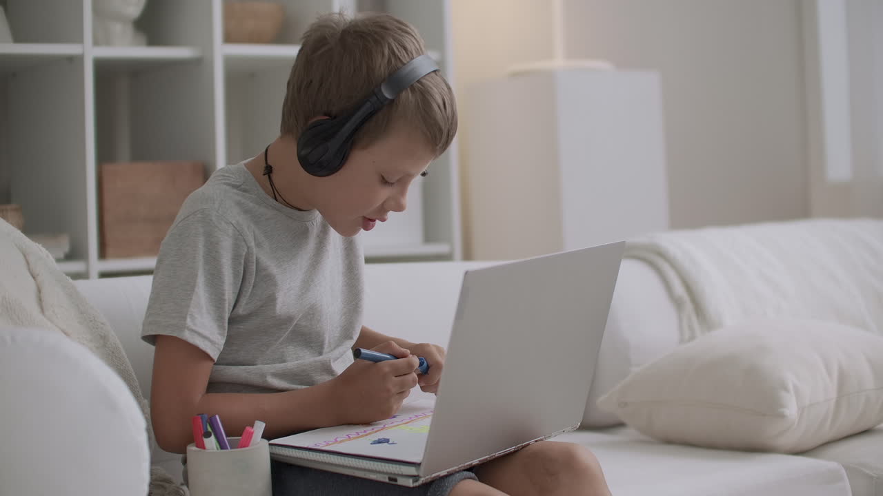 un niño contemporáneo está usando una computadora portátil y auriculares y dibujando en papel solo en la sala de estar