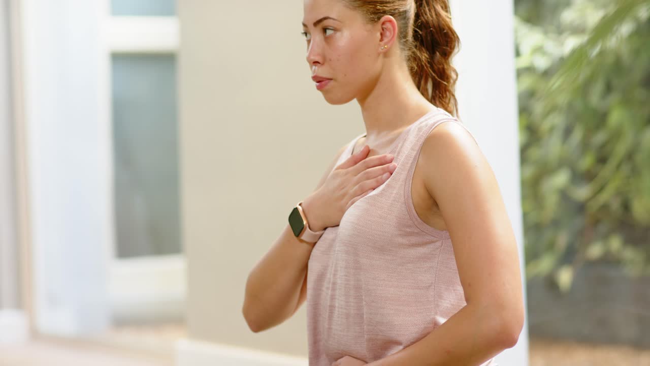 Completing breath cycles, woman rising up on green mat at home, wearing pink tank and smartwatch