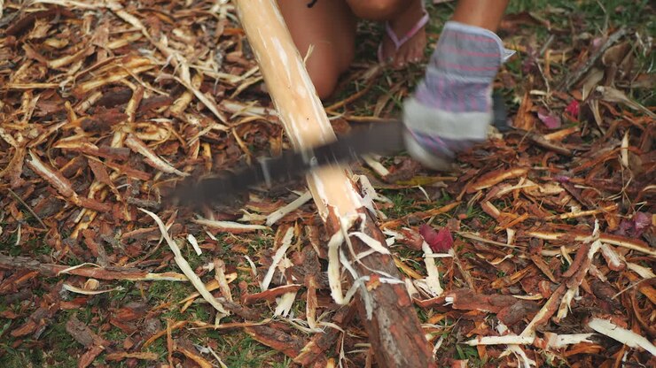 Close up of woman wearing gloves shaving bark off large stick with machete