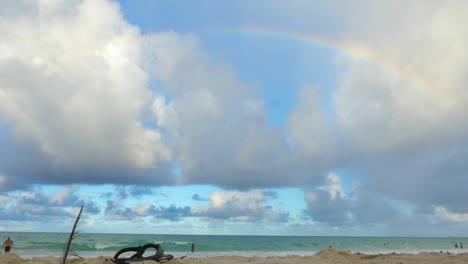 a rainbow is visible on the beach with clouds and driftwood and no people around