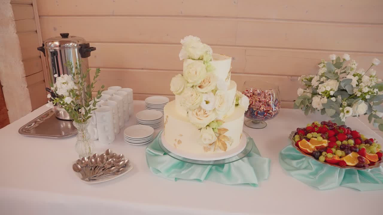 Uma mesa de bolo de casamento lindamente arranjada com decorações florais e uma travessa de frutas, ambientada contra uma parede de madeira. A cena apresenta luz suave e natural acentuando a montagem elegante.