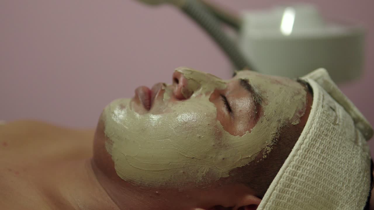 Latin mexican man guy relaxing enjoy skin care therapy massage at spa wellness center with clay mask on face costumer client