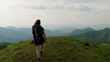 Woman-backpacker-hikes-up-to-grassy-overlook-down-on-lush-tropical-misty-valley,-rear-view-static