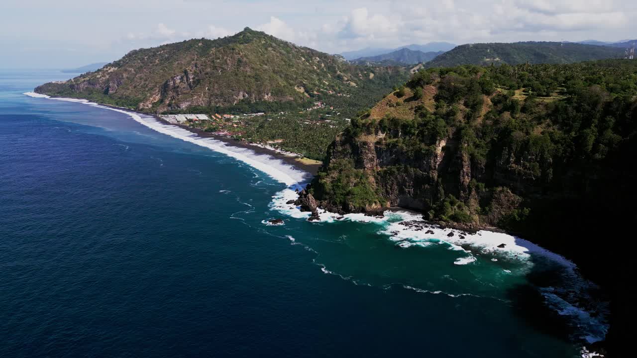 Drone view of Karangasem Bali’s pristine black sand beach where gentle ocean waves meet rolling green hills under a bright sunny sky, creating a perfect coastal paradise scene