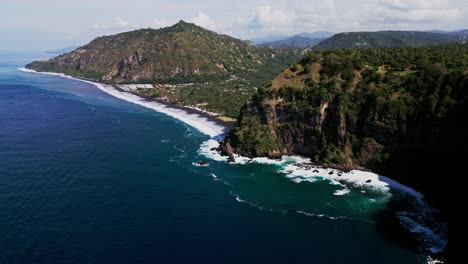 drone view of karangasem bali’s pristine black sand beach where gentle ocean waves meet rolling green hills under a bright sunny sky, creating a perfect coastal paradise scene