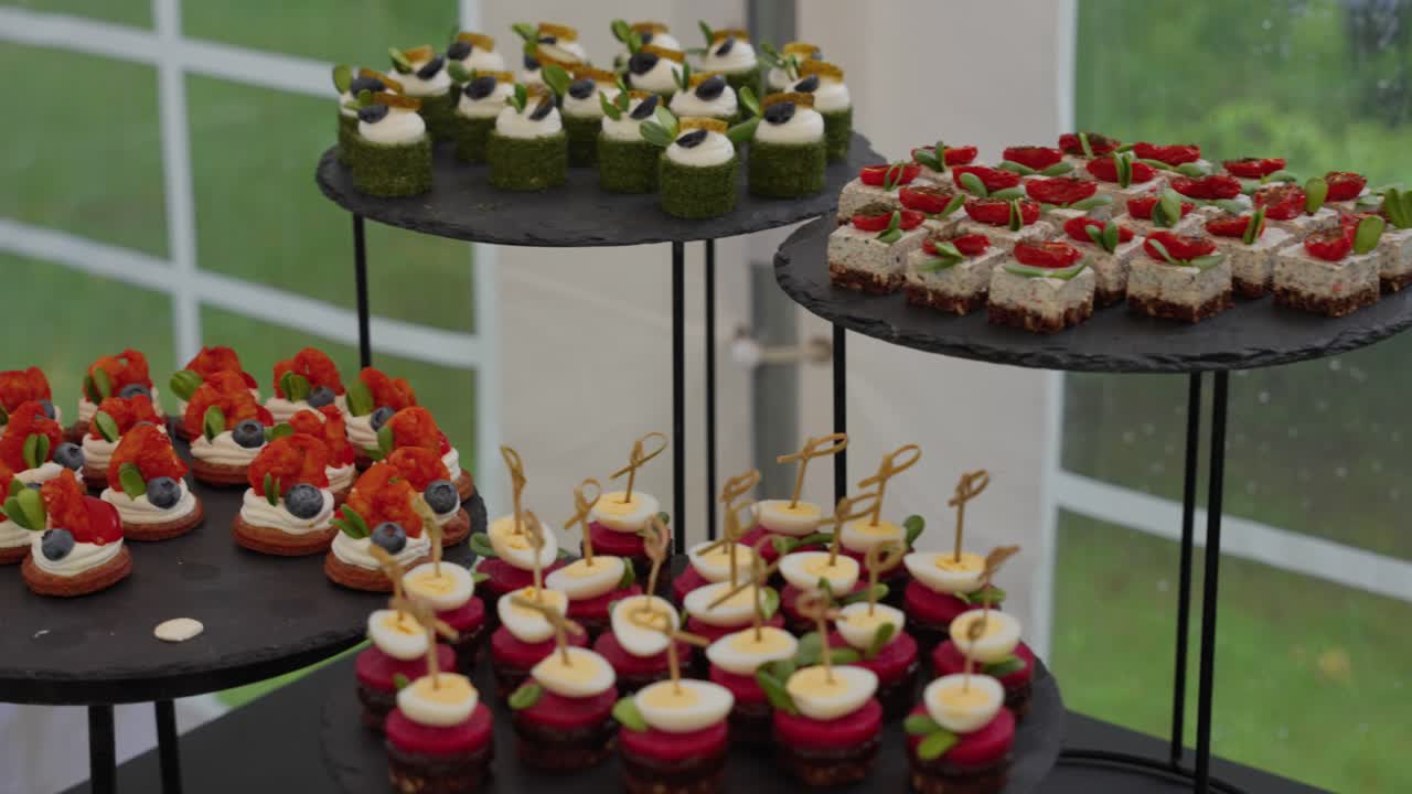 Raffinata composizione di dessert gourmet disposti su vassoi neri a più livelli durante un evento all'aperto. Catturata con illuminazione soffusa, le prelibatezze colorate creano un'esperienza culinaria visivamente accattivante.