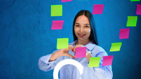 brunette with long hair turns to camera holding hands in a shape of heart. beautiful girl at the glass board with stickers put in a shape of heart.