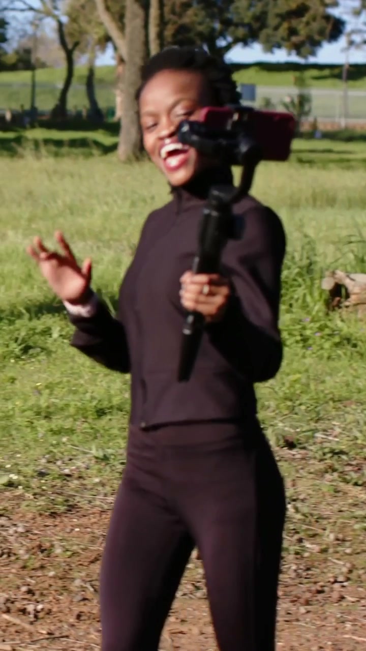 Video verticale: Telecamera in movimento, donna con scarpe da ginnastica viola che cammina su un sentiero del parco facendo vlogging con un gimbal rosso.