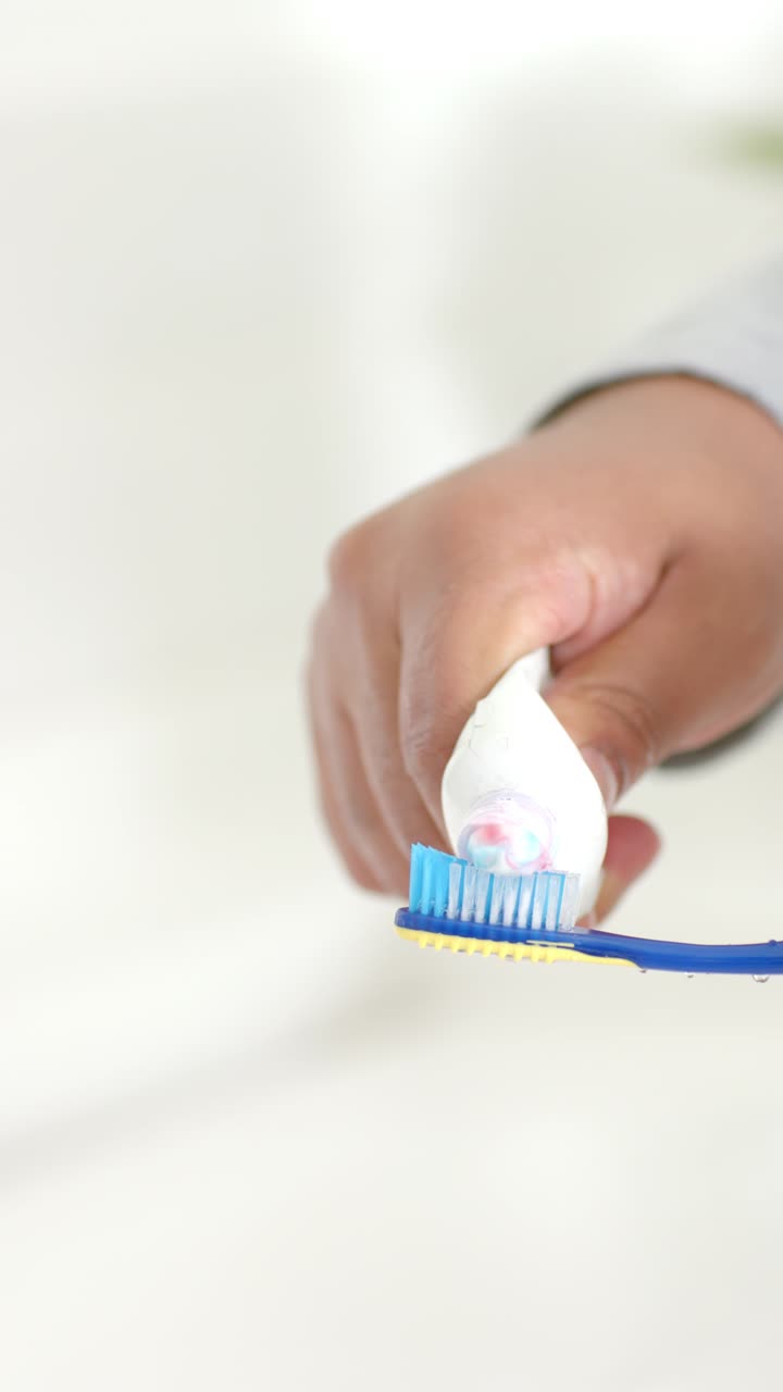 Man squeezing tube dispensing pink-white paste onto blue toothbrush at vanity preparing to brush