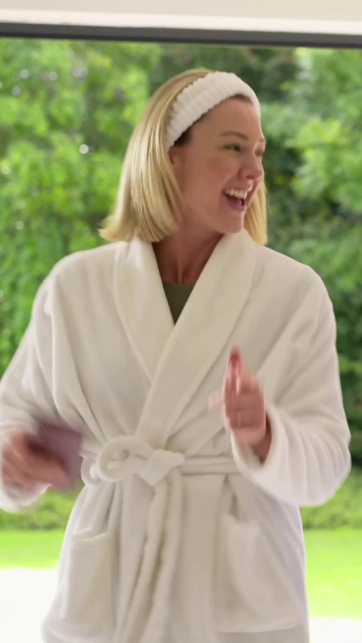 Vertical video: Lifting phone, woman at home in robe and headband turning, gesturing to greet guest