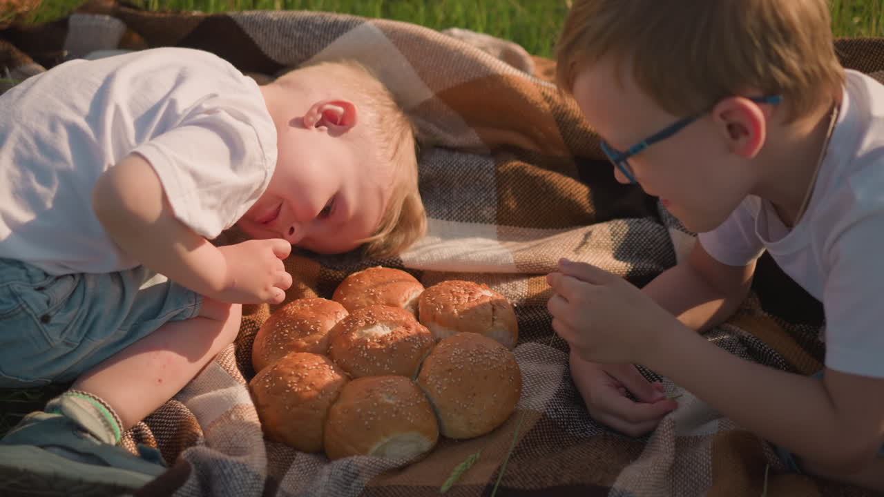 Two young boys wearing white shirts are lying on a checkered scarf in a grassy field, enjoying pieces of freshly baked bread.The peaceful outdoor setting highlights a simple serene moment of childhood