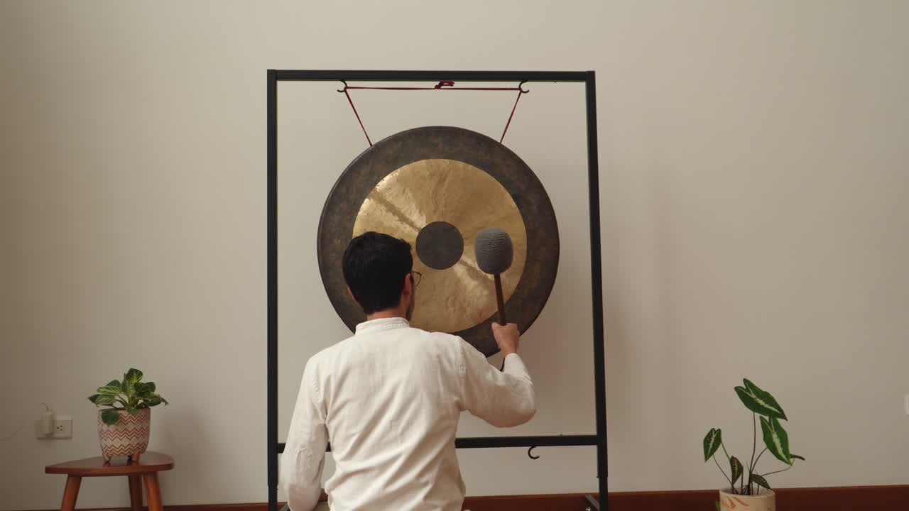 Rear view of a man striking a large symphonic gong with a soft mallet in a calm minimalist indoor studio with plants—ideal for wellness, meditation, sound healing and therapy concepts
