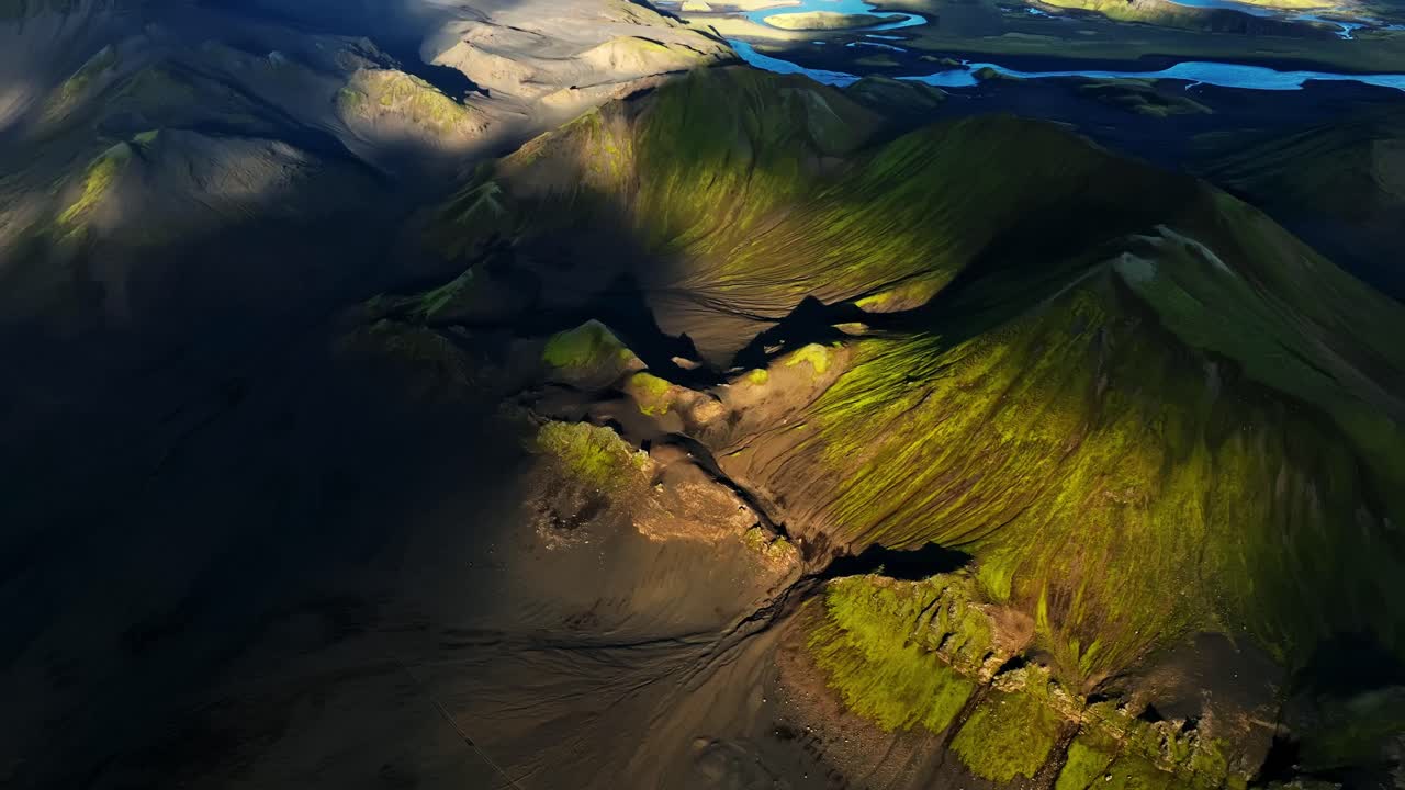 Aerial shot drifting above Iceland’s shadowed volcanic slopes, revealing sharp moss-lined ridges, eroded channels and glowing patches of light across a dramatic highland basin