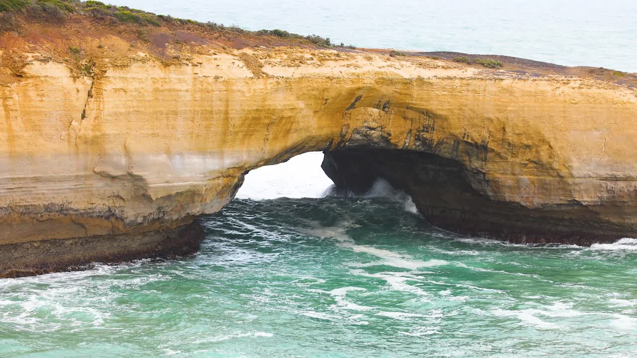 Dynamic ocean waves crash against the iconic London Bridge rock formation under soft daylight at Port Campbell, Australia