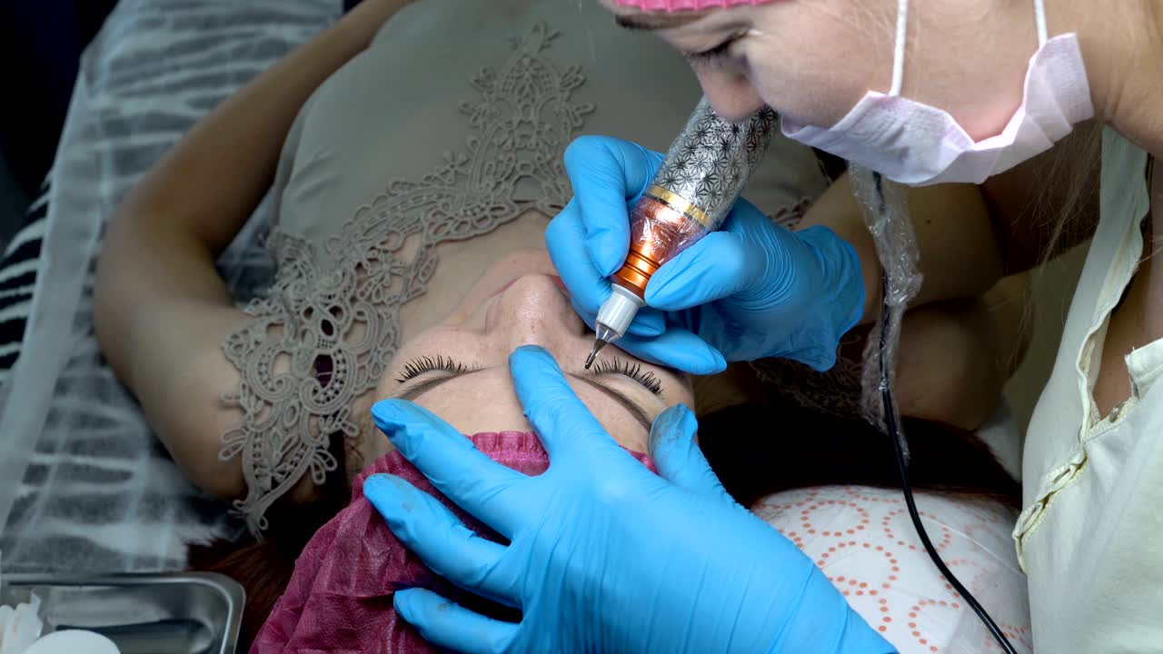 A young girl doing permanent makeup eyebrows in a beauty salon. The beautician puts black paint on her eyebrows with a needle. Microblading procedure. Close up. The view from the top. 4K, 25 fps.