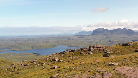 Cinematic-drone-shot-of-Stac-Pollaidh-in-the-Northwest-Highlands-of-Scotland,-passing-over-rocky-mountain-tops-to-reveal-bodies-of-water