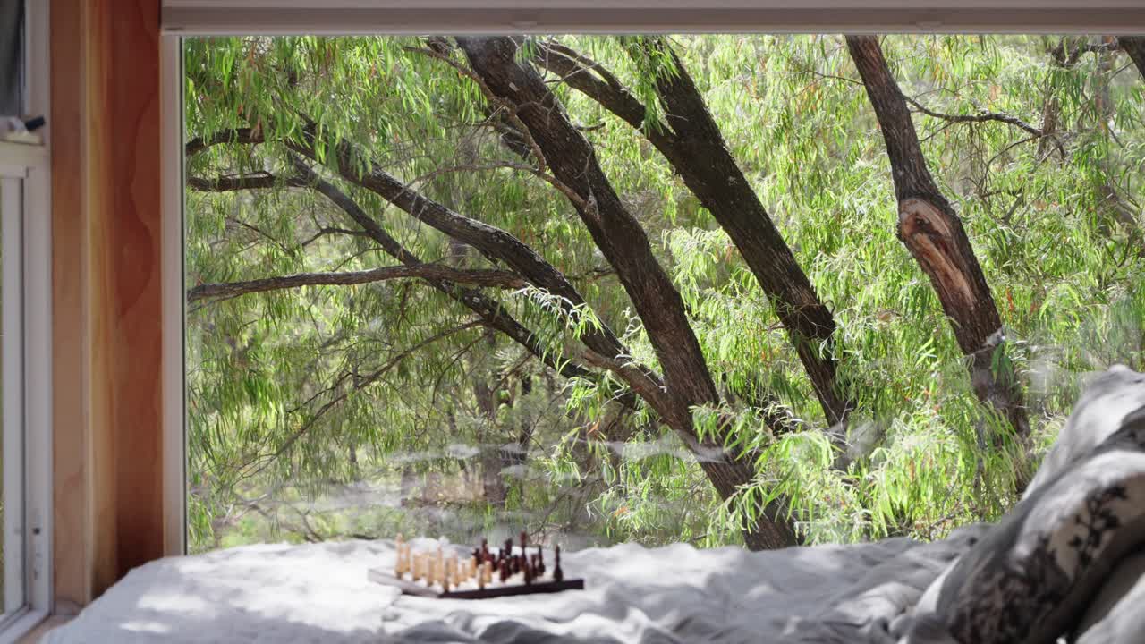 체스 보드는 숲으로 큰 창문을 가진 작은 오두막 침대의 꼭대기에 앉아 있습니다.