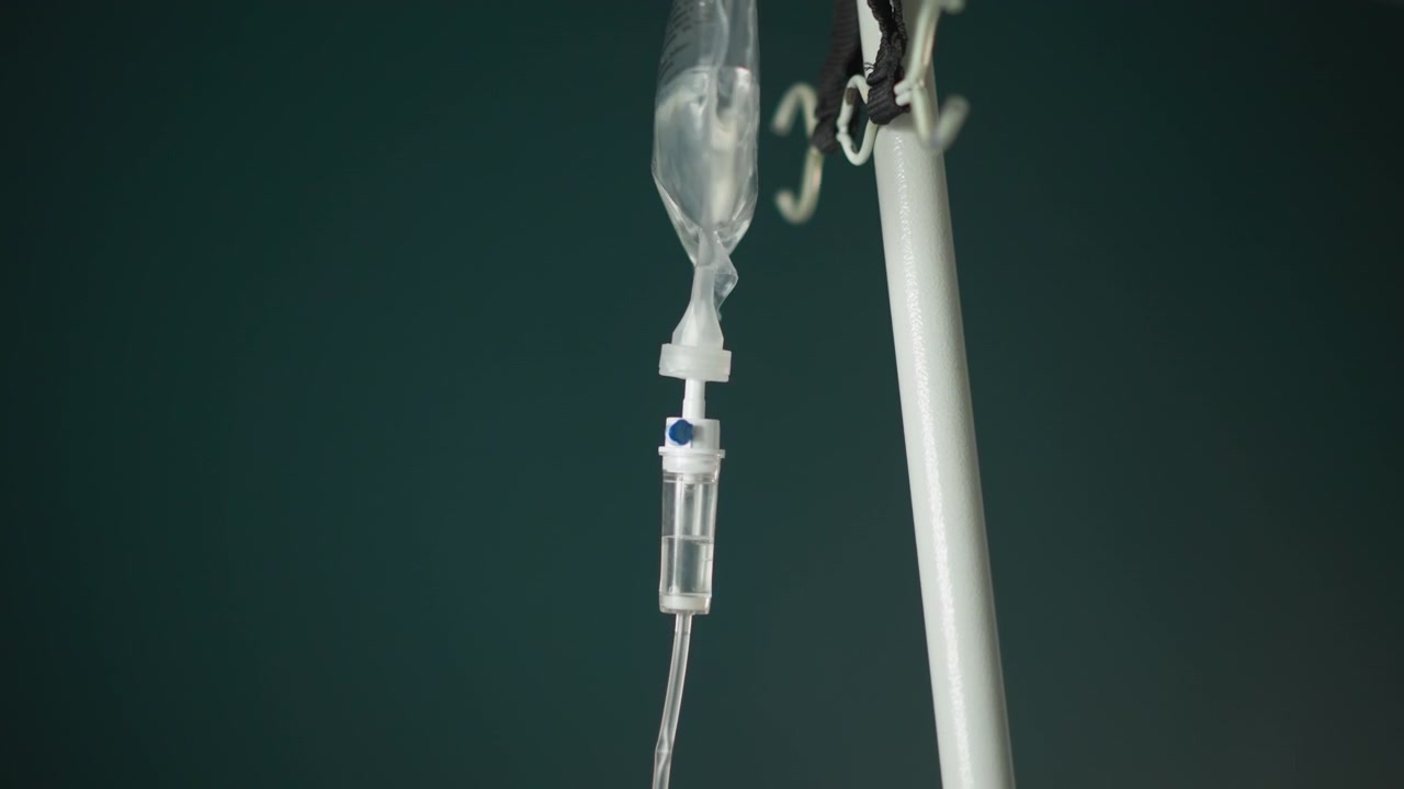 Main gantée ajustant une perfusion intraveineuse, le bout du doigt manipulant le régulateur de débit sur un tube transparent sous une poche de solution saline, contre un mur clinique sarcelle. Gros plan sur le gant stérile et l'ajustement précis, un moment rassurant.